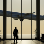 Silhouette of a traveller standing in an airport terminal, watching a passenger airplane take off through large glass windows.