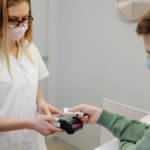 A healthcare worker wearing a face mask and white uniform hands a health card to a masked patient at a clinic reception desk.