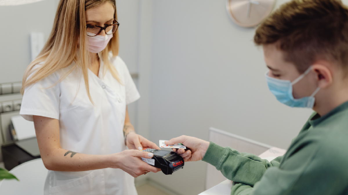 A healthcare worker wearing a face mask and white uniform hands a health card to a masked patient at a clinic reception desk.