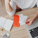An overhead view of a person sitting at a desk holding receipts beside a laptop, notebook, cash, and stationery, illustrating bill payments, expense tracking, or managing household finances.