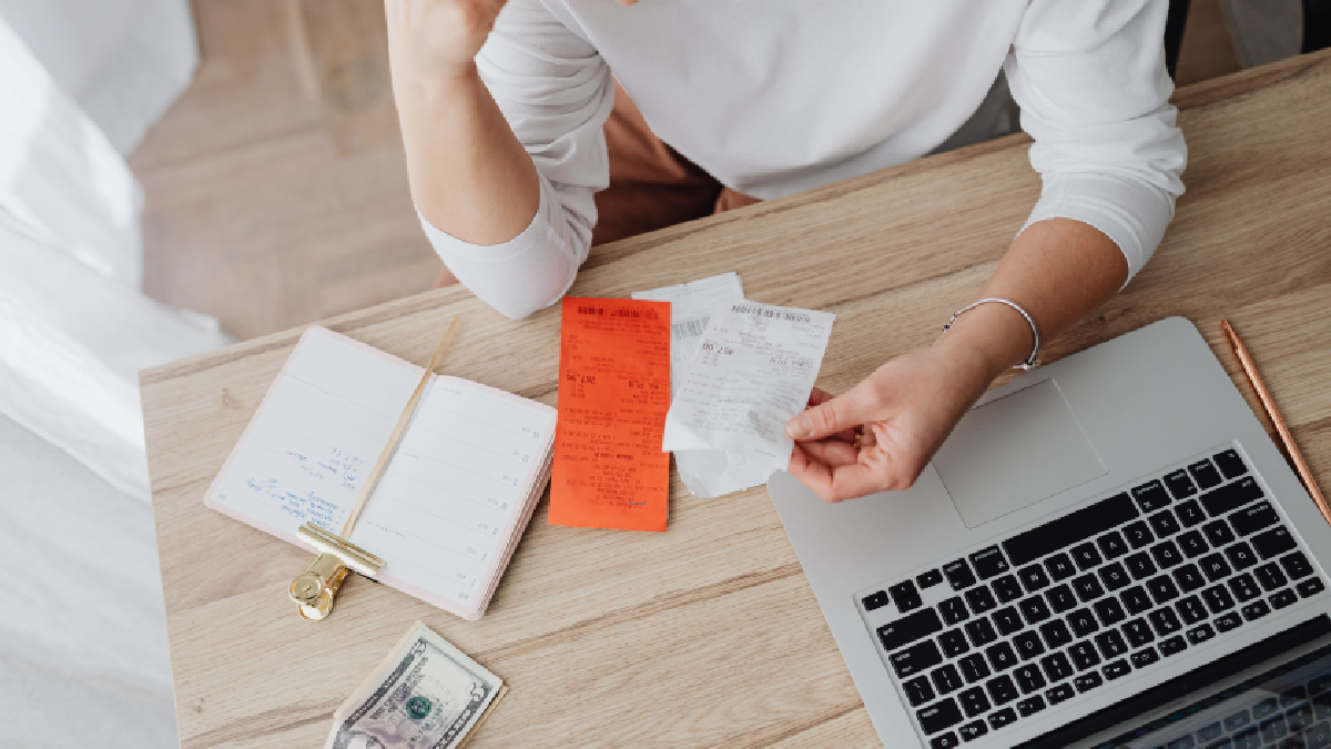An overhead view of a person sitting at a desk holding receipts beside a laptop, notebook, cash, and stationery, illustrating bill payments, expense tracking, or managing household finances.