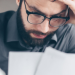 A worried man wearing glasses reviews financial documents while holding his forehead, conveying stress over loans, debt, or financial obligations.
