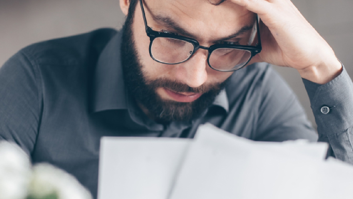 A worried man wearing glasses reviews financial documents while holding his forehead, conveying stress over loans, debt, or financial obligations.