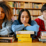 Three international students resting their arms on stacks of textbooks. In a library, looking directly at the camera.