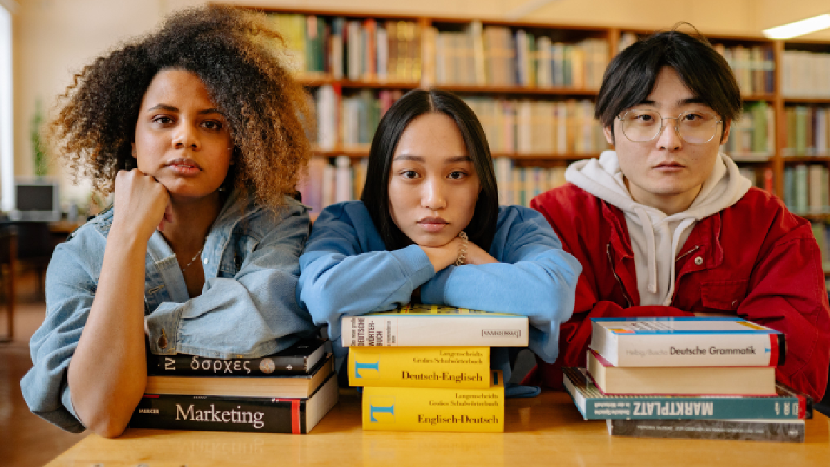 Three international students resting their arms on stacks of textbooks. In a library, looking directly at the camera.